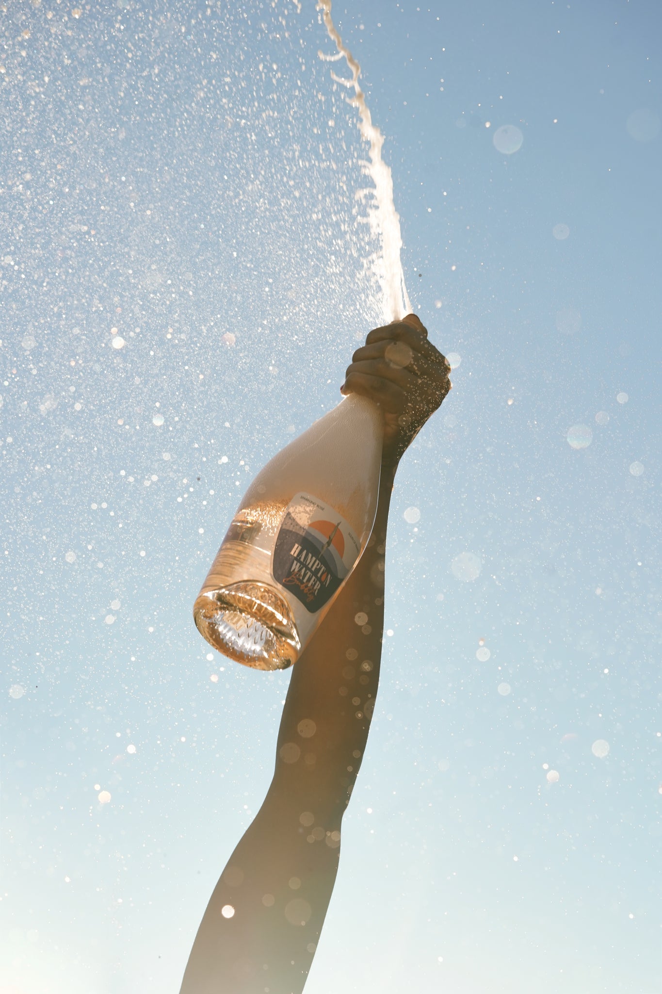 Arm holding up a bottle of Hampton Water Bubbly as t bubbles skyward